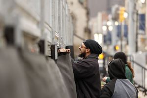 A person in a beanie and dark jacket uses a spray can on a wall in an urban street setting.