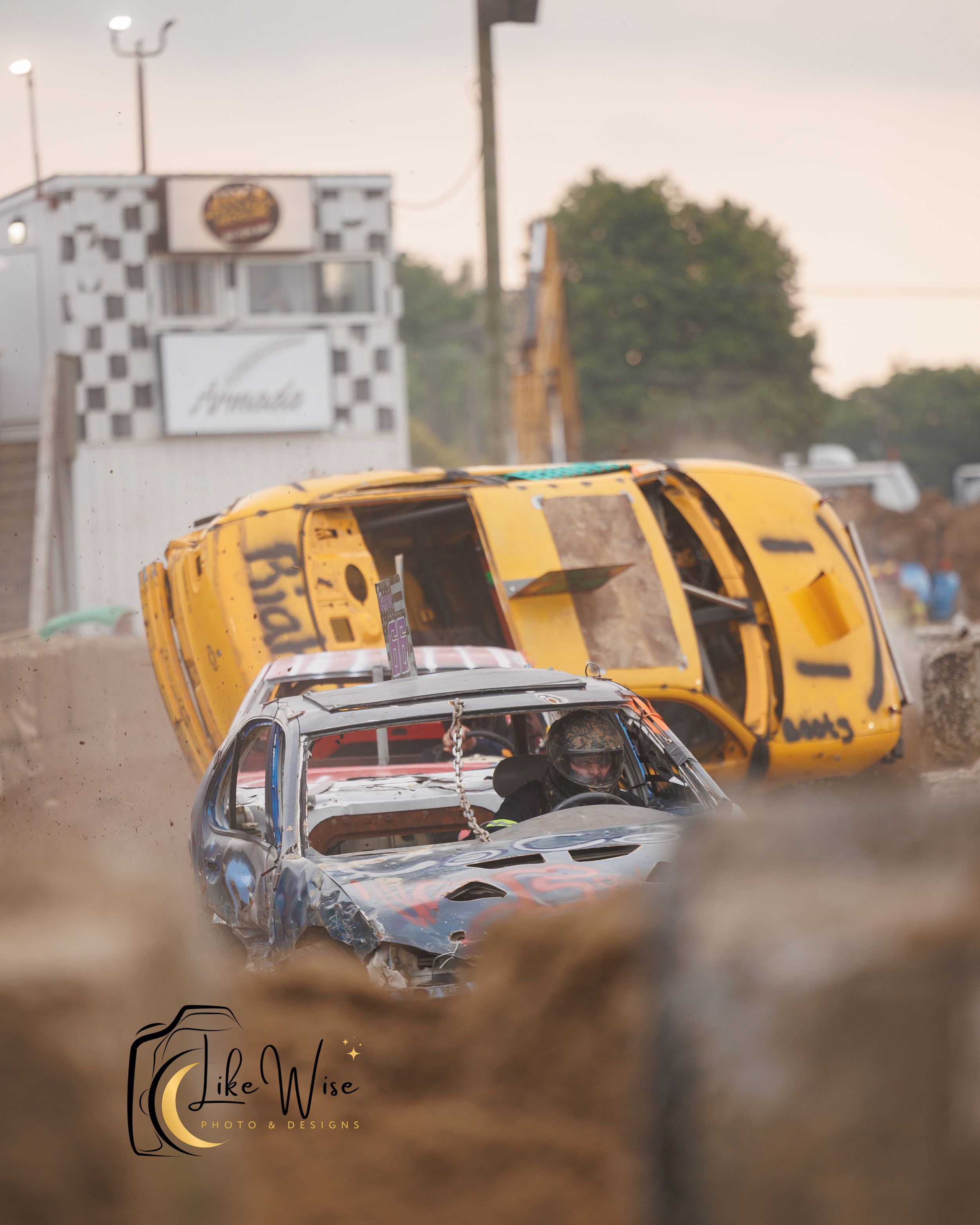 Two demolition derby cars collide violently at a race track, with one yellow vehicle flipping over in a cloud of dust.