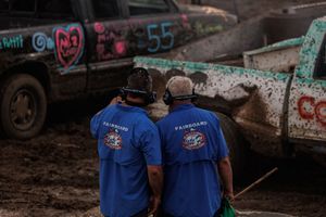 Two people in matching blue t-shirts stand watching a demolition derby event with muddy cars in the background.