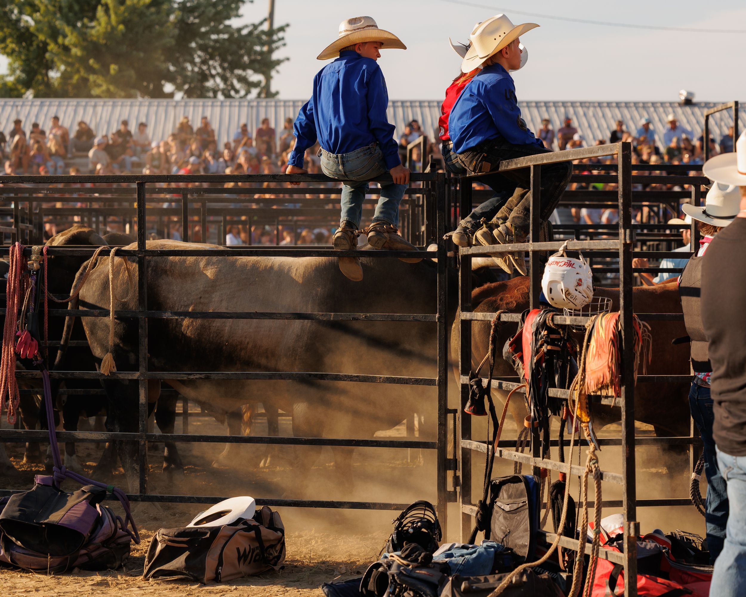 Cowboys in western attire watch from wooden railings at a rodeo event as riders compete in the arena.