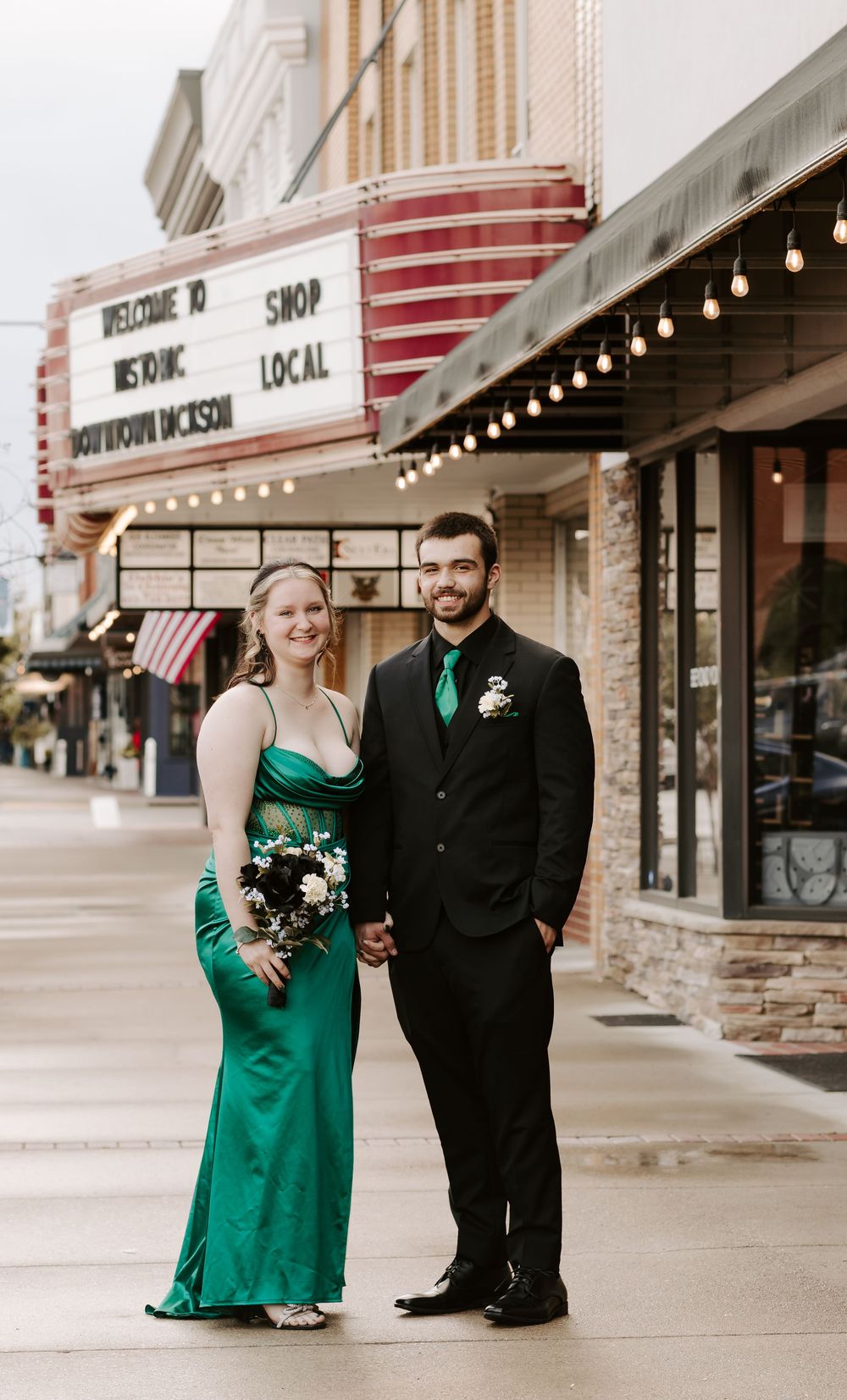 A couple in formal attire poses outside a vintage movie theater with a classic marquee and string lights.