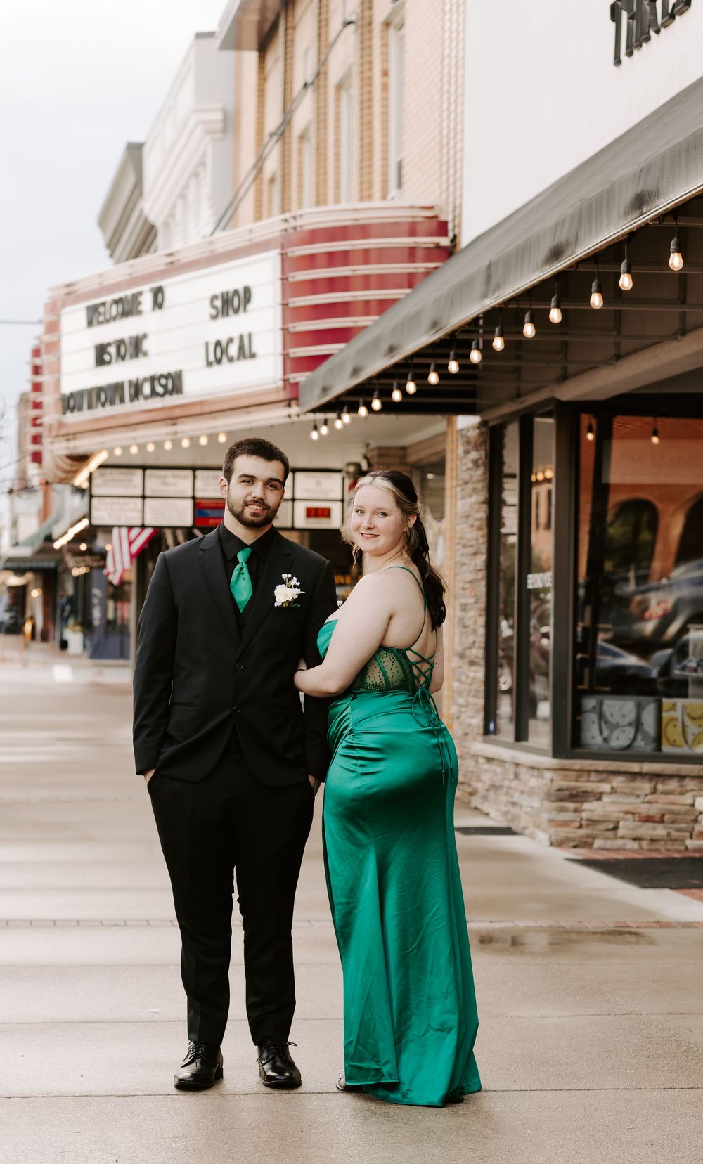 A couple poses together in front of downtown storefronts, with the person in black and their partner wearing emerald green.