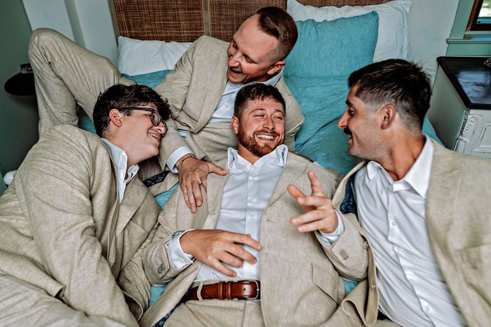A group of friends in beige suits laugh together while lounging on a bed with turquoise pillows.