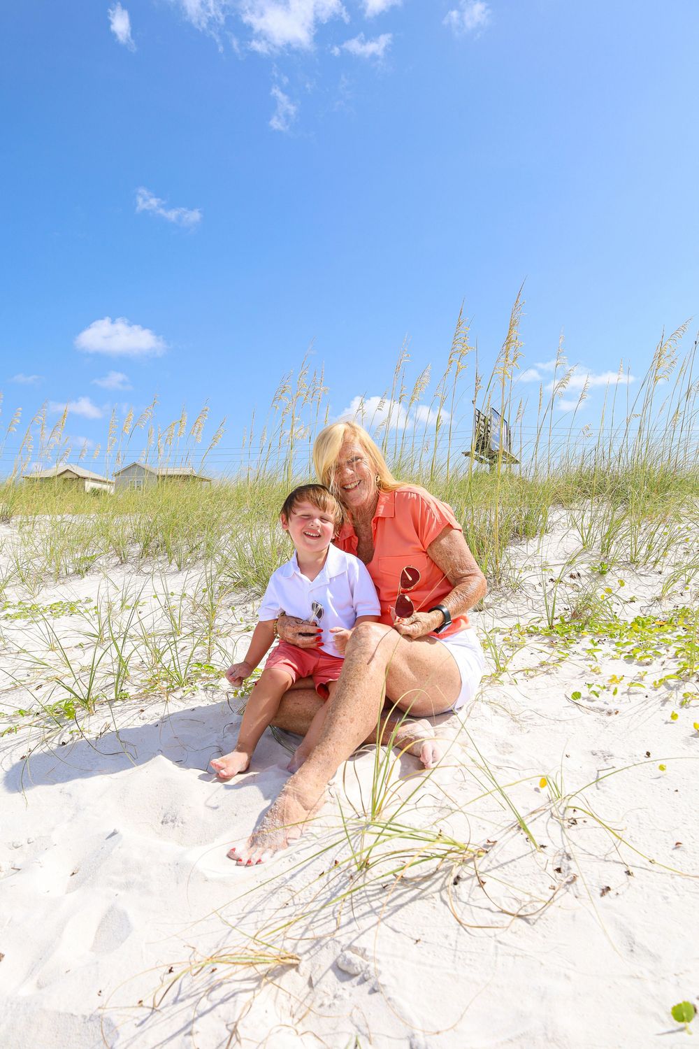 Two people sit together on white sandy beach dunes with sea oats and blue sky on a sunny summer day.
