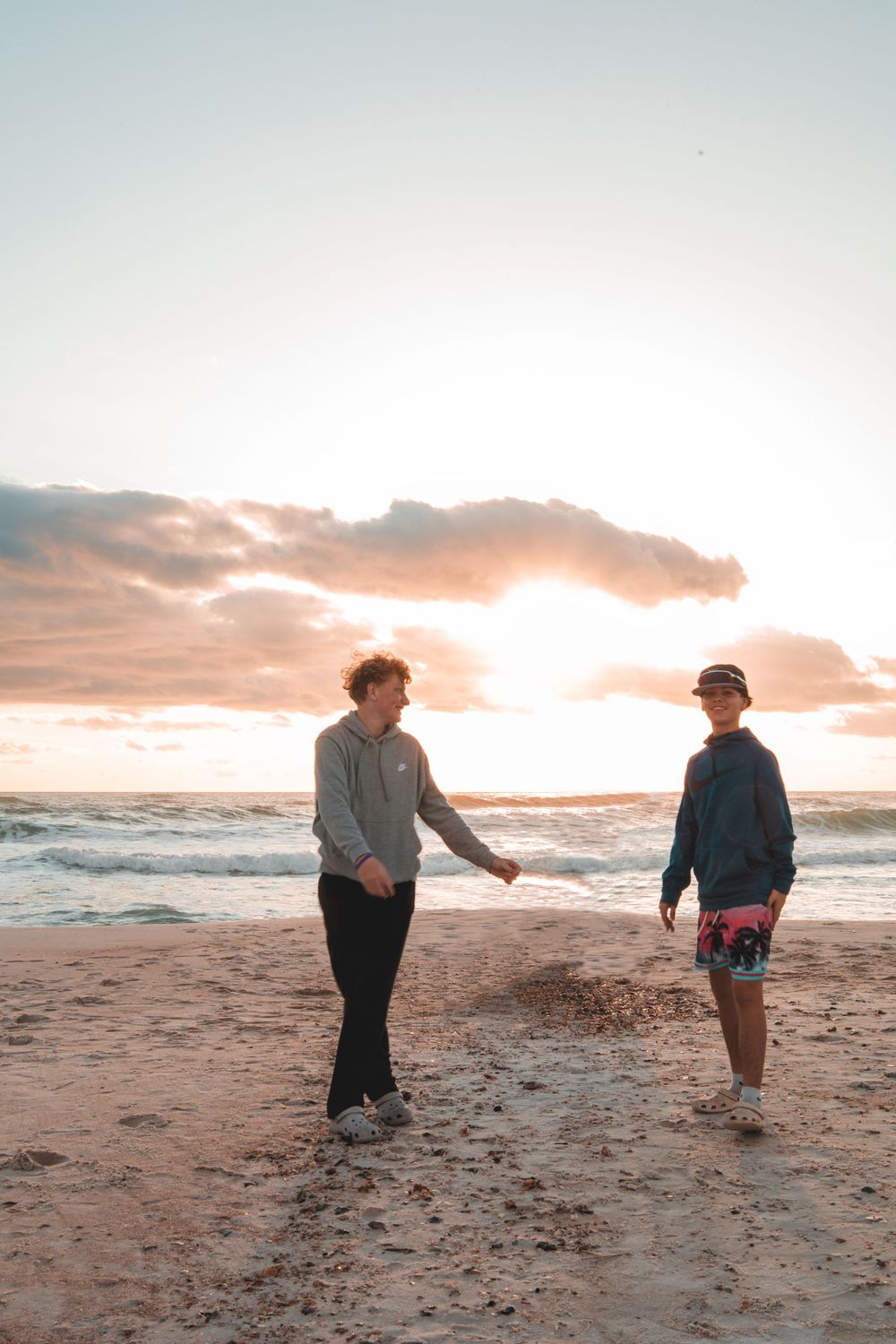 Two people walk along a sandy beach at sunset while waves crash against the shore.
