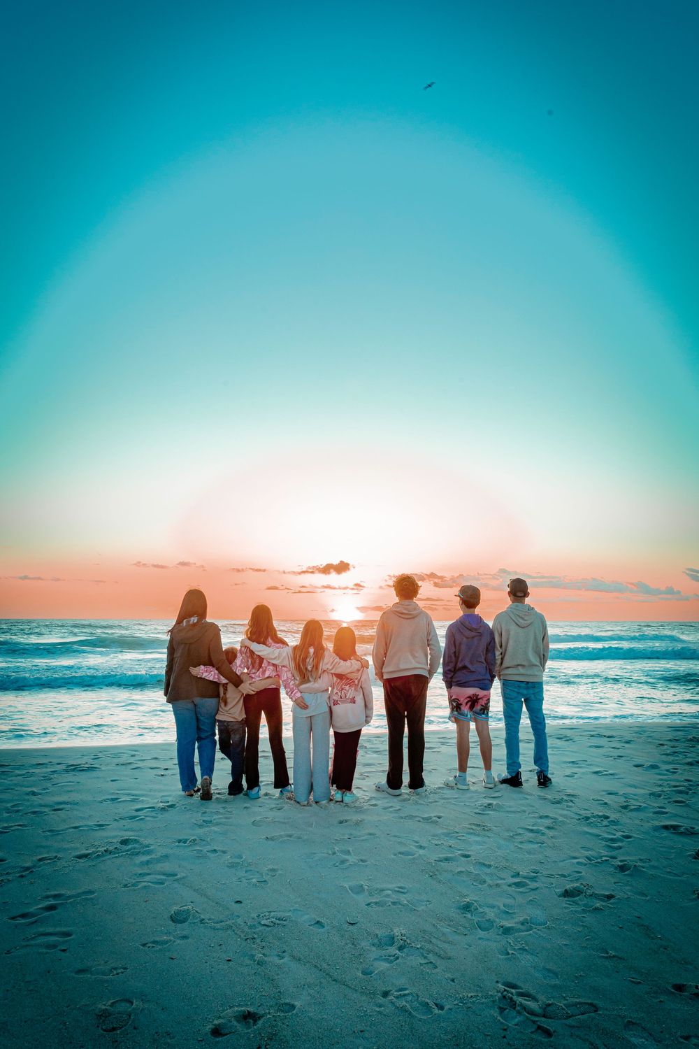 Group of people standing together on beach at sunset watching ocean waves with turquoise sky and orange horizon.