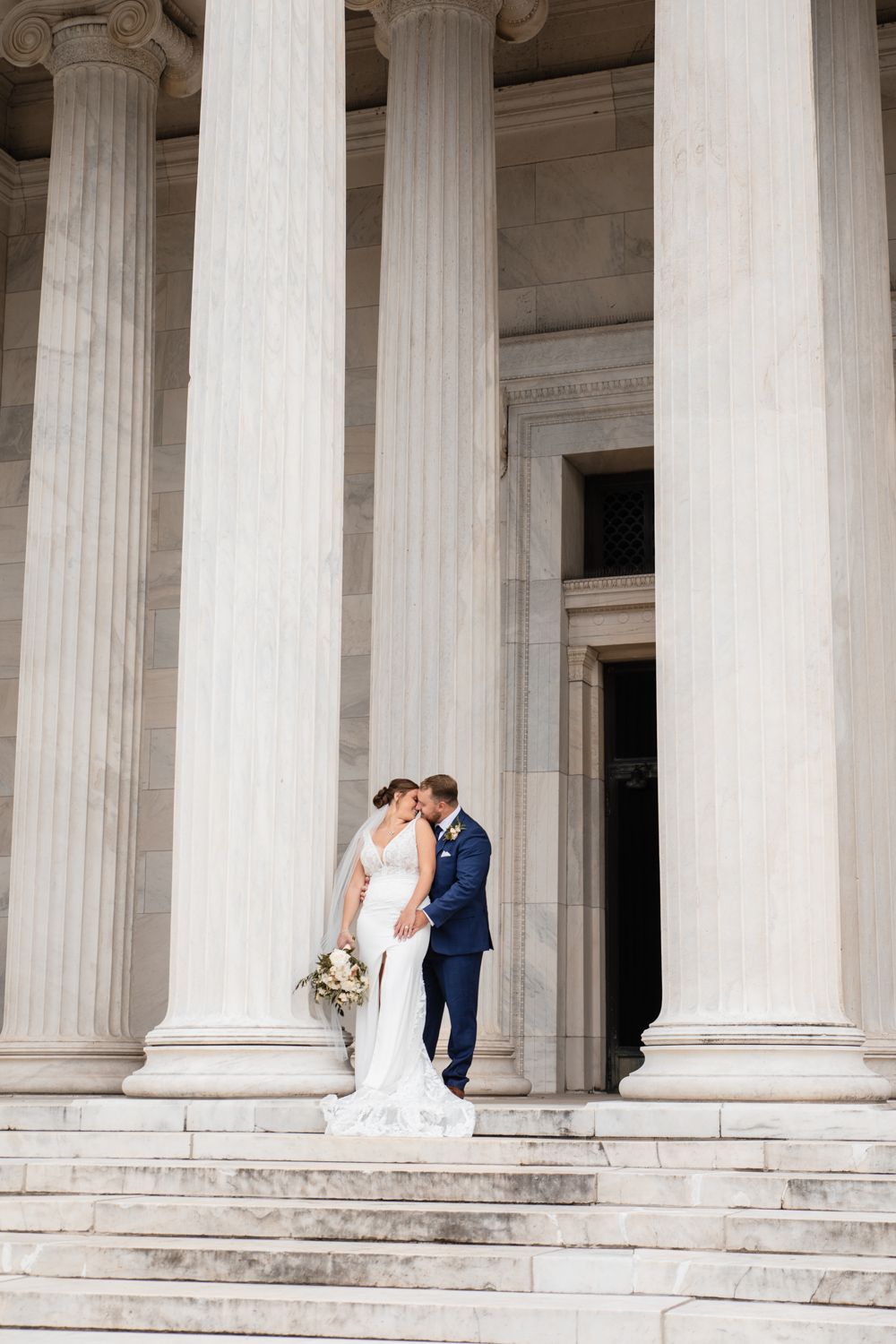 A couple stands intimately between grand white marble columns on the steps of a classical building.