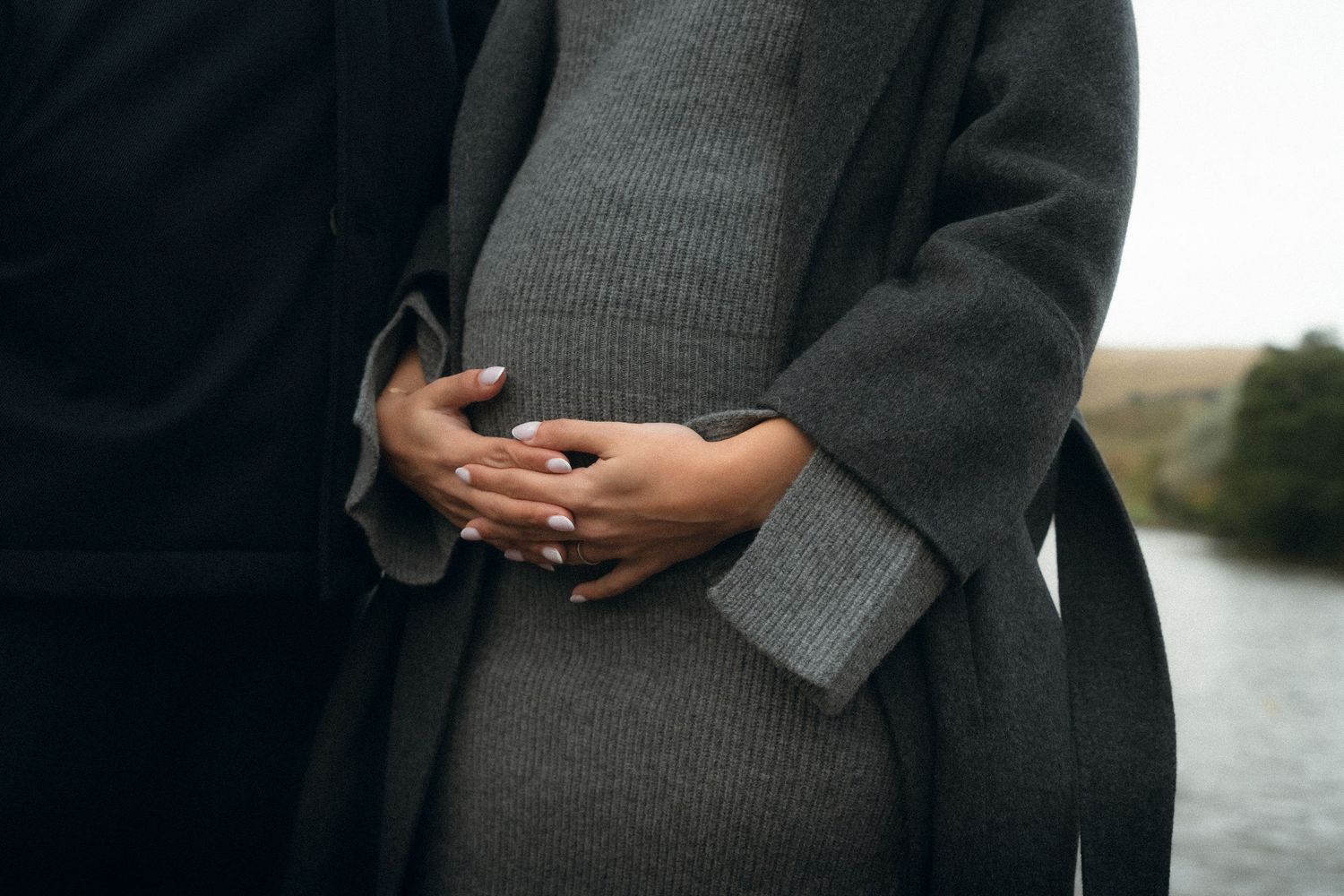 A series of maternity photos showing a pregnant woman in a black dress and gray cardigan by a waterfront.