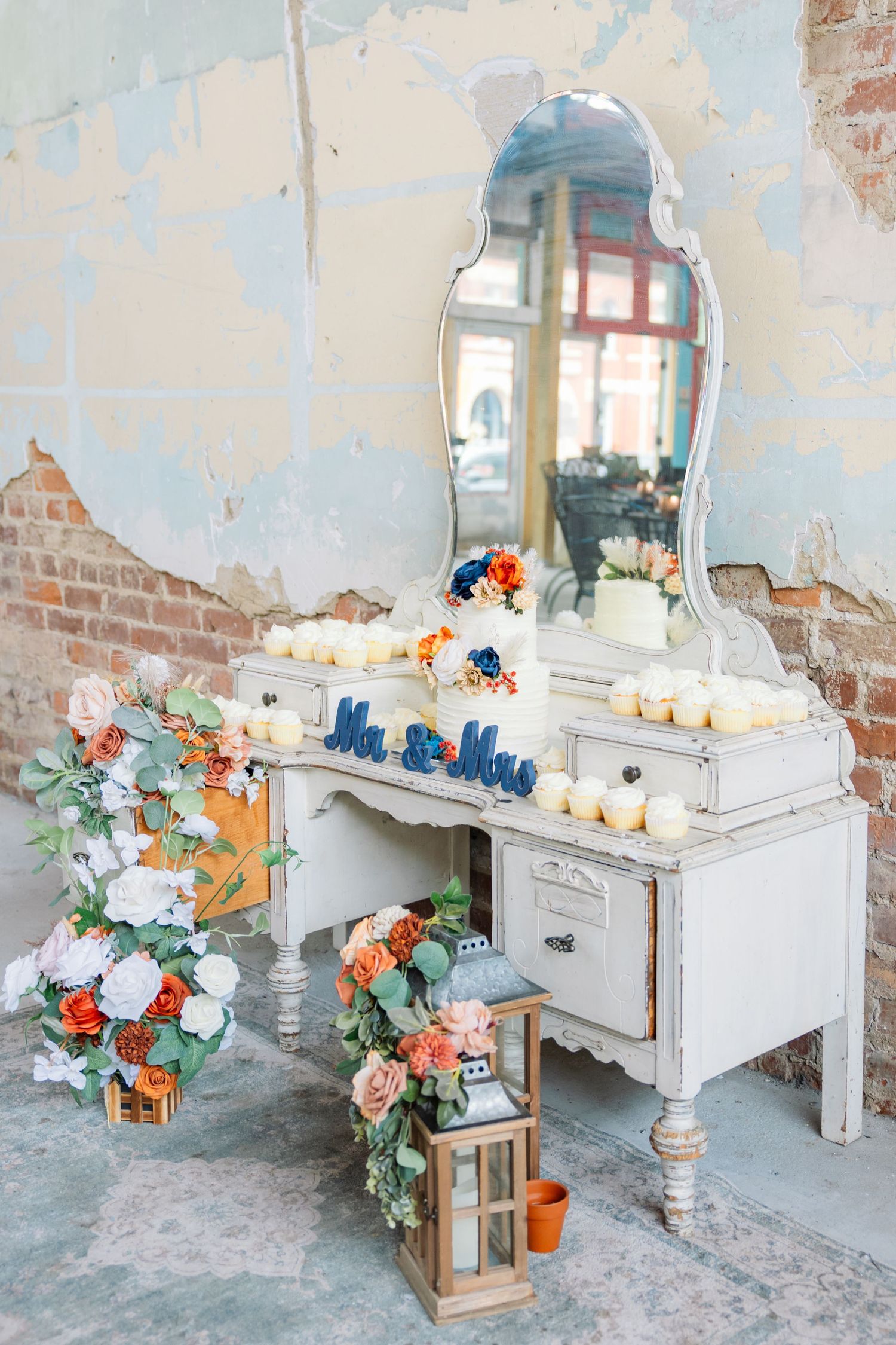 Vintage white vanity decorated with colorful floral arrangements, lanterns, and desserts in a rustic brick setting.