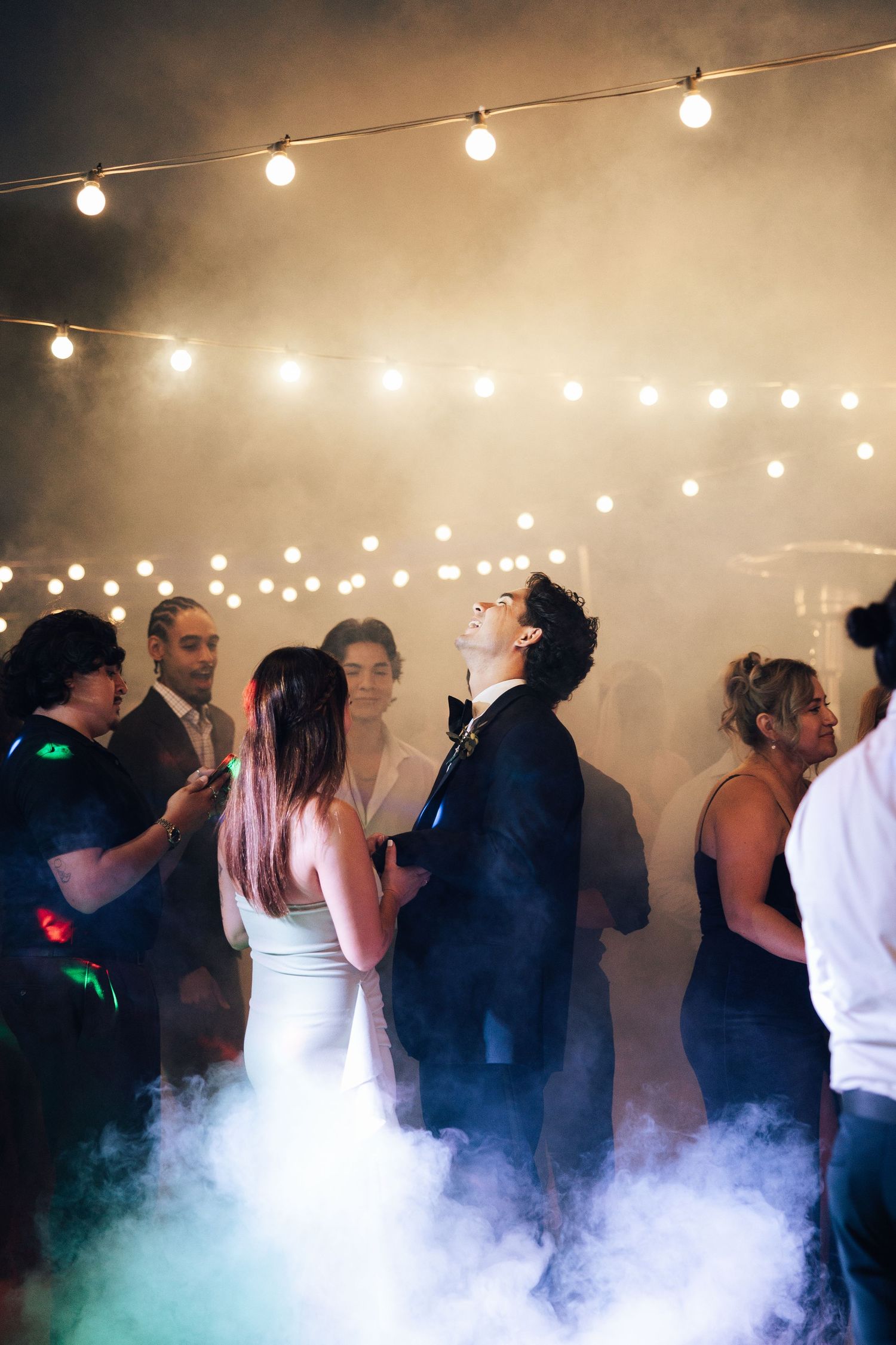 Party guests dance on a foggy dance floor under string lights creating a dreamy atmosphere at an indoor event.