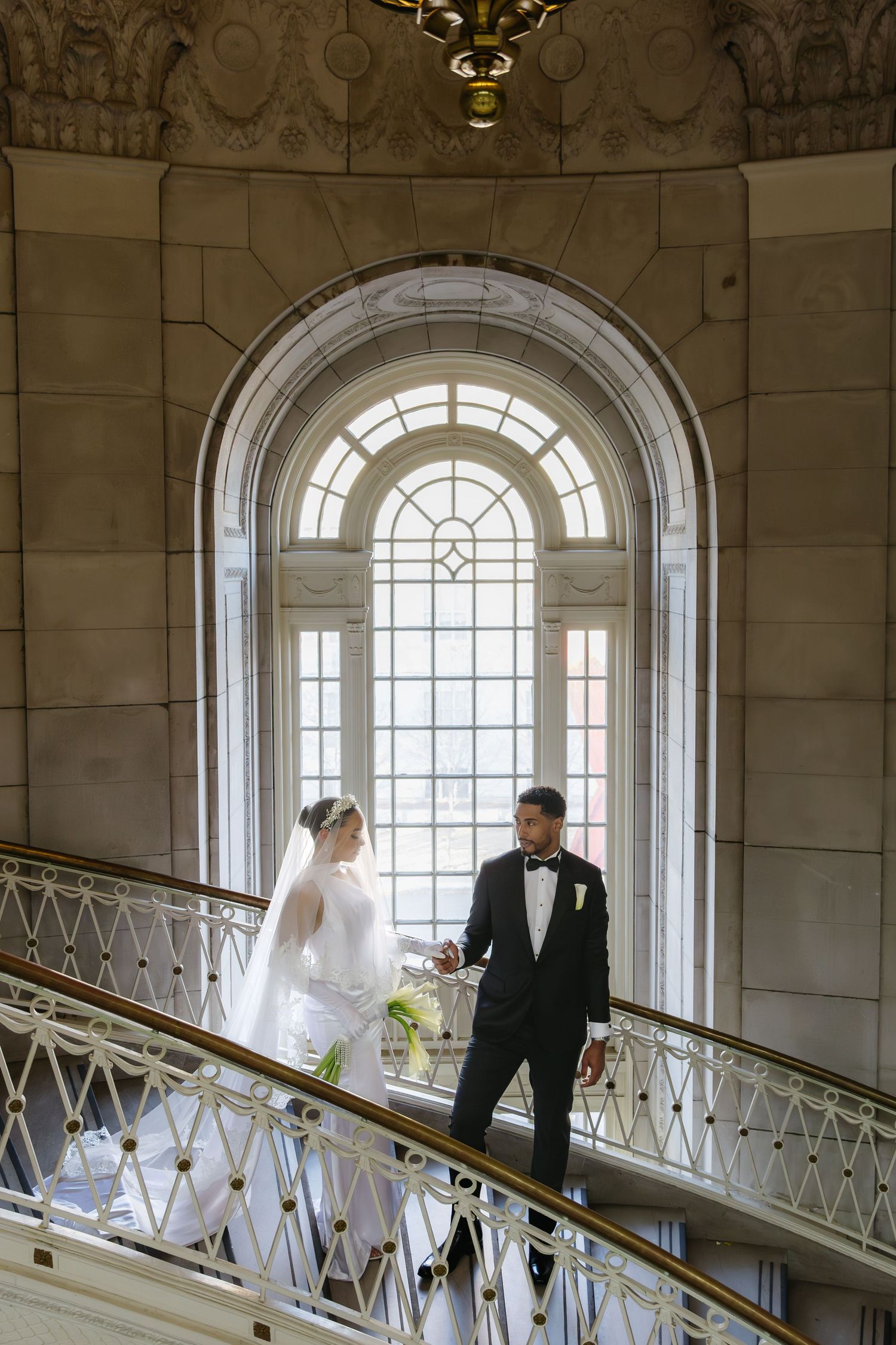 A couple stands on an elegant curved staircase with a grand arched window and ornate railing in a historic building.