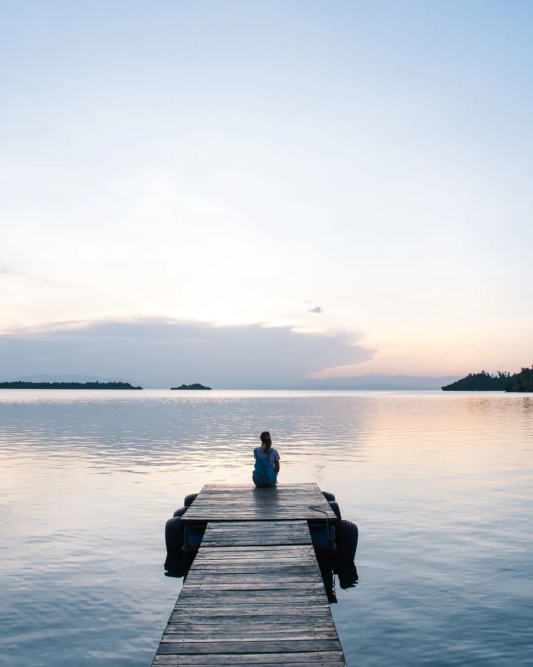 Person standing at the end of a wooden pier looking out over a calm lake at sunrise.