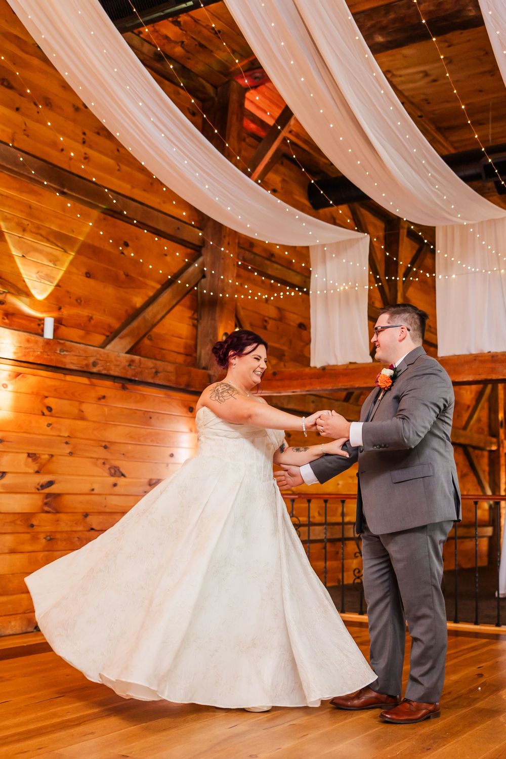 Couple dancing in a rustic wooden barn venue decorated with white fabric draping and twinkling string lights.