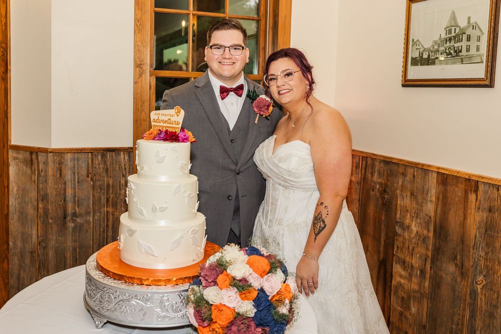 Three-tiered white wedding cake decorated with orange and navy blue floral accents at a rustic reception.