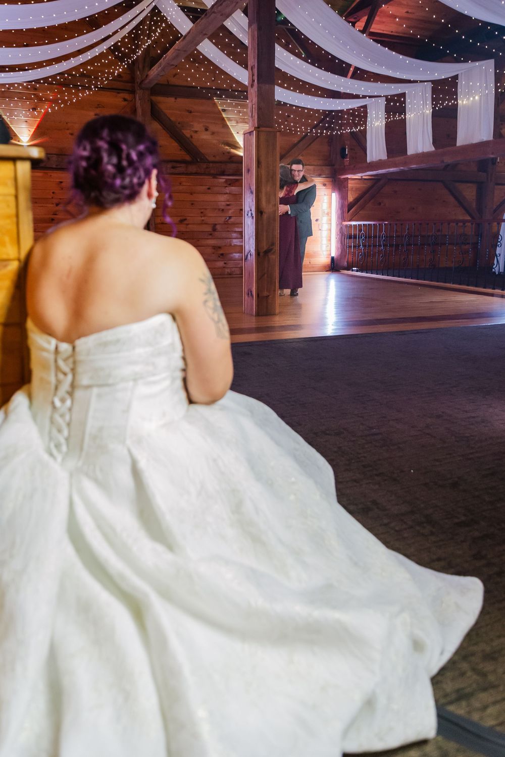Bride in flowing white wedding dress twirls on rustic wooden dance floor under string lights at barn reception.