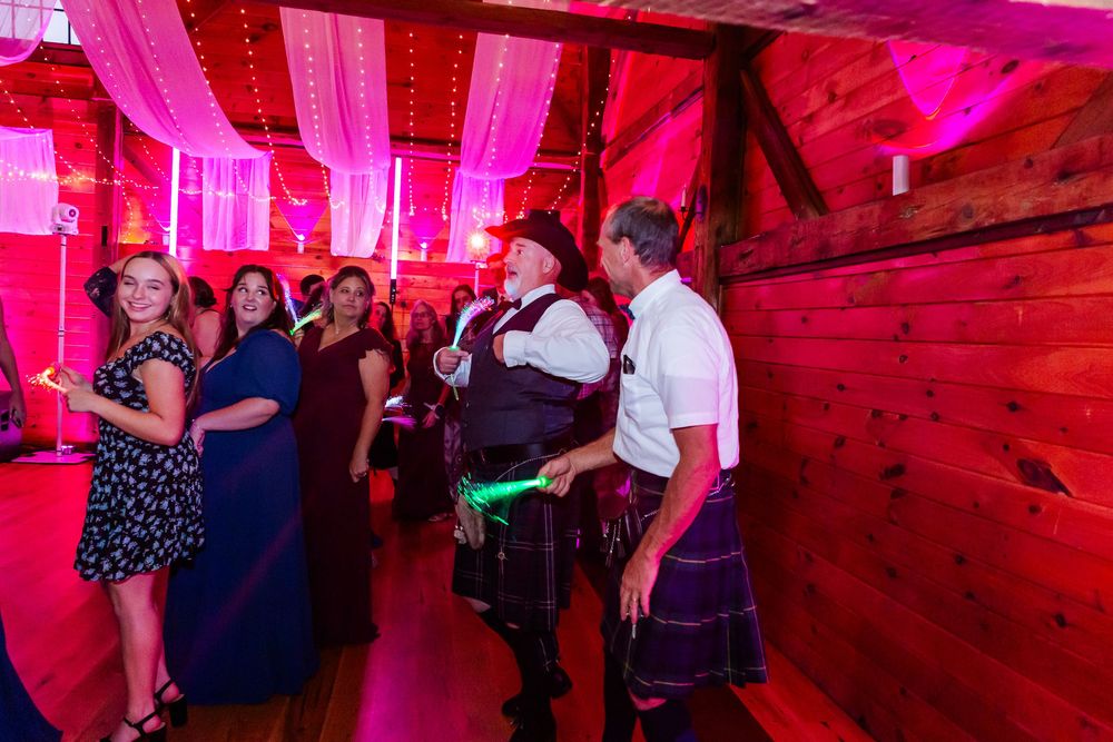 Wedding guests dancing and celebrating on a dance floor illuminated by vibrant pink lighting in a rustic venue.