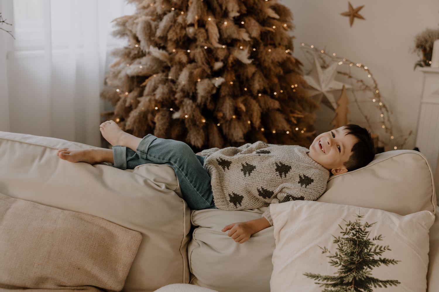 Small child in beige dress stands smiling in front of a fireplace decorated with holiday lights and greenery.