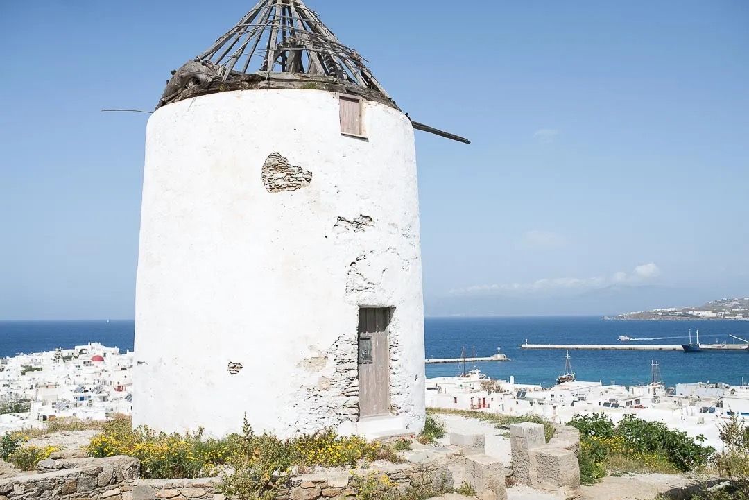 A classic white Greek windmill on a rocky hillside with sea views behind.
