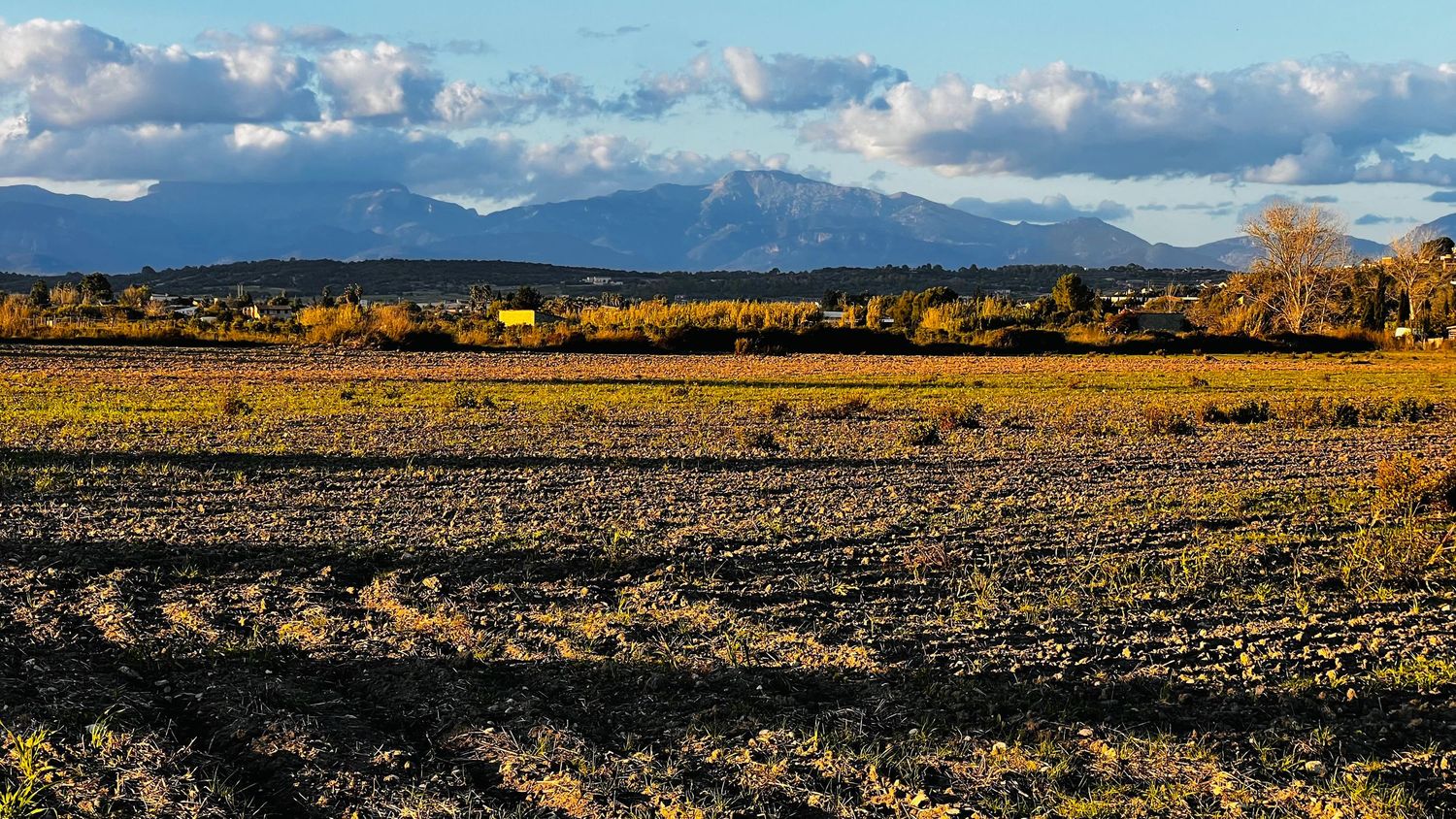 A panoramic mountain landscape with snow-capped peaks and rolling hills under a dramatic cloudy sky at sunset.