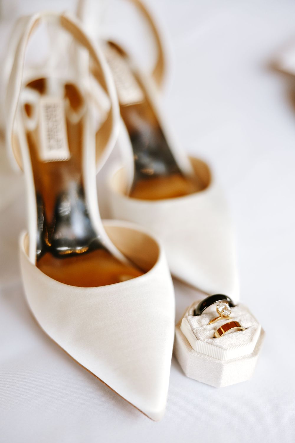 Elegant white satin bridal heels with pointed toes displayed next to a small ring box on light background.