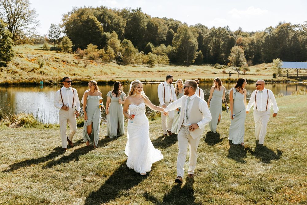 A group in white attire walks through a sun-drenched meadow during a rustic outdoor celebration.