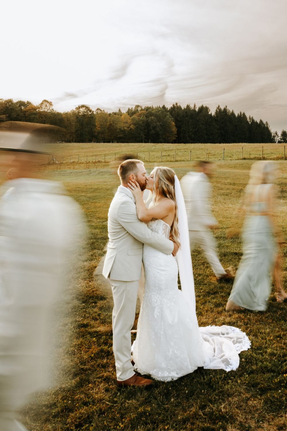 A couple shares a romantic kiss in a golden field at sunset with blurred ethereal figures in the foreground.