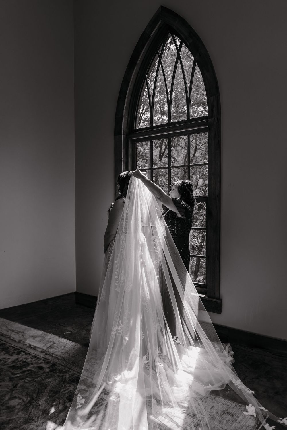 A flowing white wedding veil cascades in front of an arched window creating a dramatic silhouette in black and white.