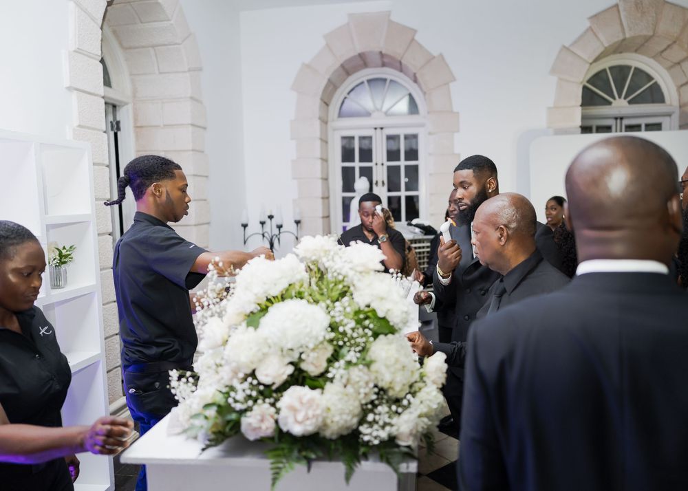 Wedding party members gather in an elegant white ballroom with arched windows and chandeliers.