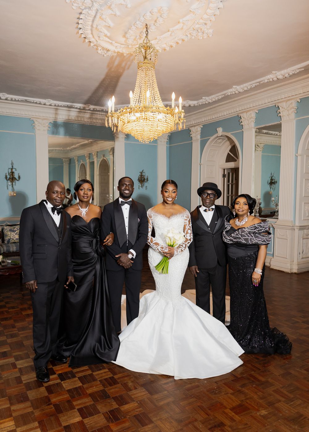 Wedding party celebrates in grand ballroom with blue walls and ornate chandelier.