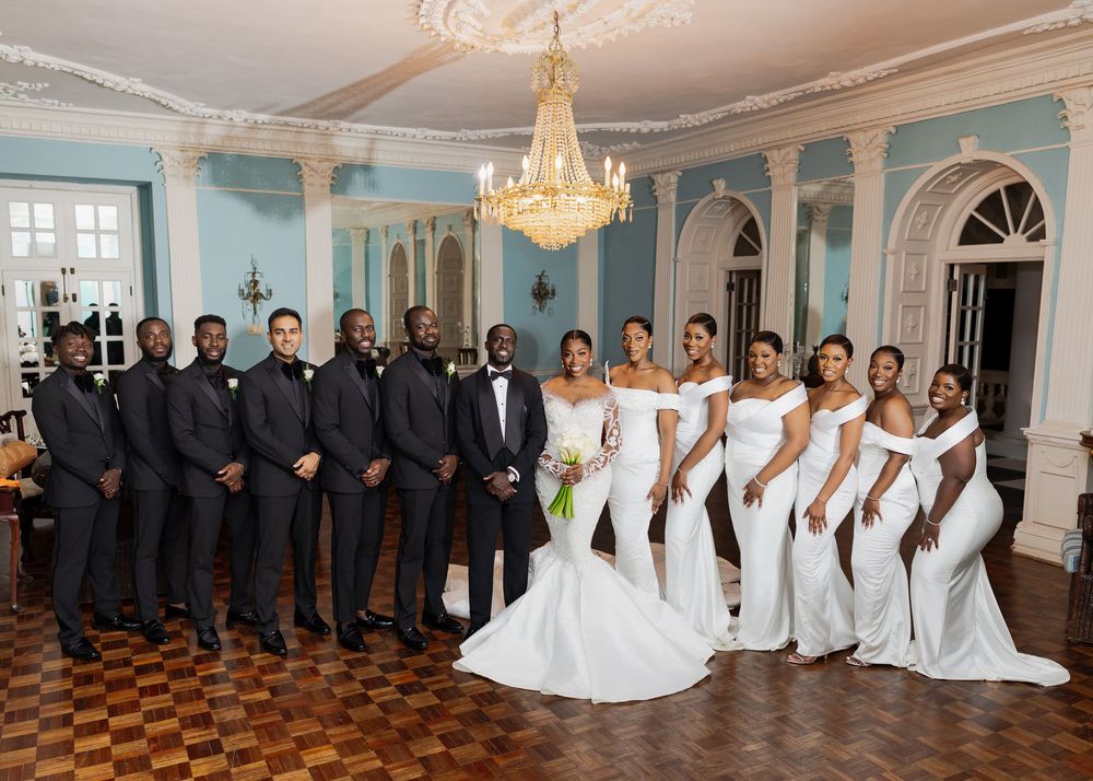 Wedding party poses in formal attire inside a grand ballroom with blue walls and checkerboard flooring.
