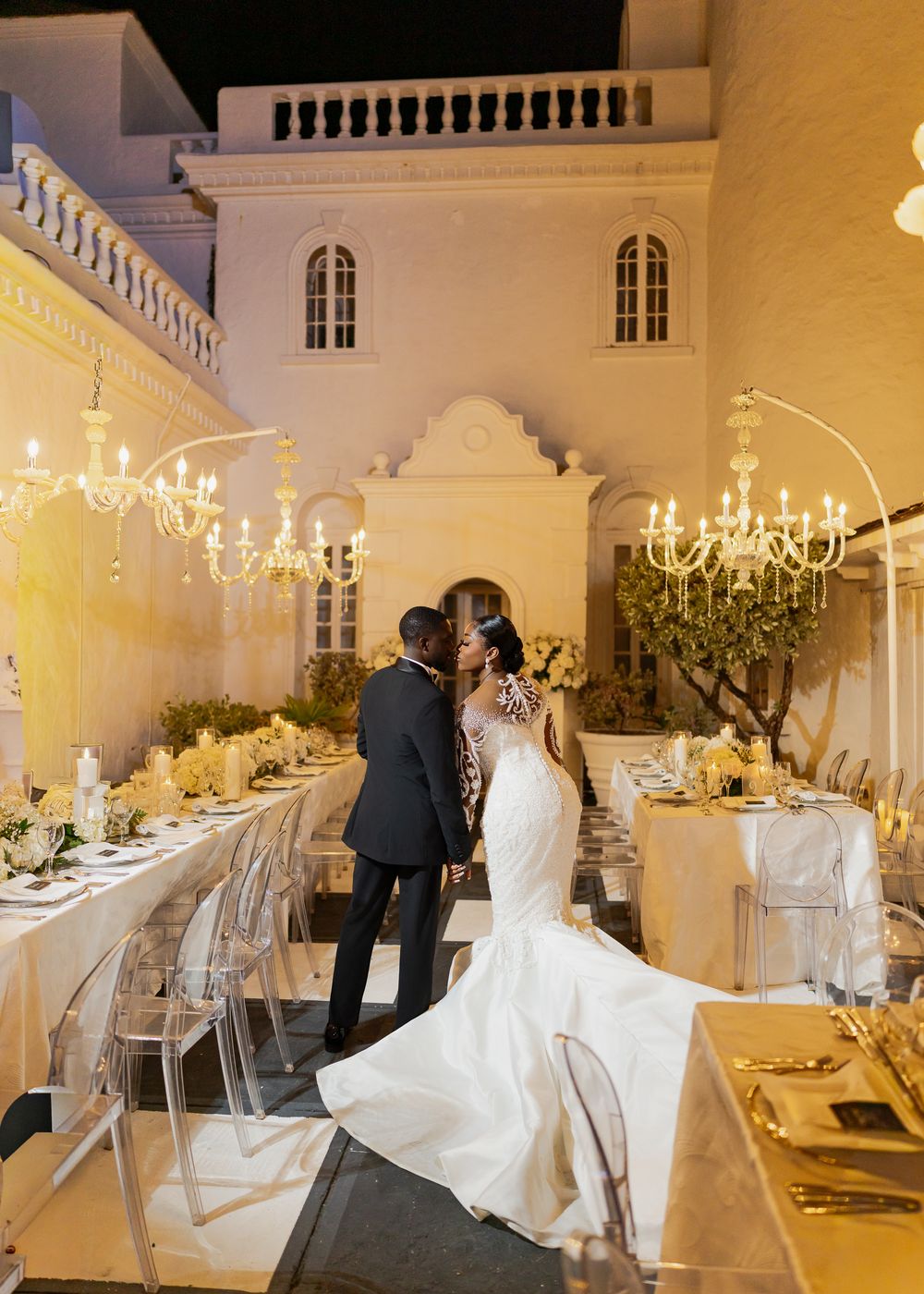 Elegant wedding reception venue with crystal chandeliers, ghost chairs, and white floral centerpieces on long dining tables.