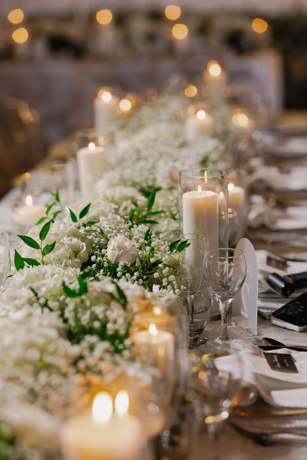 Elegant wedding table setting with white floral centerpieces, glowing candles, and green foliage creating a romantic ambiance.