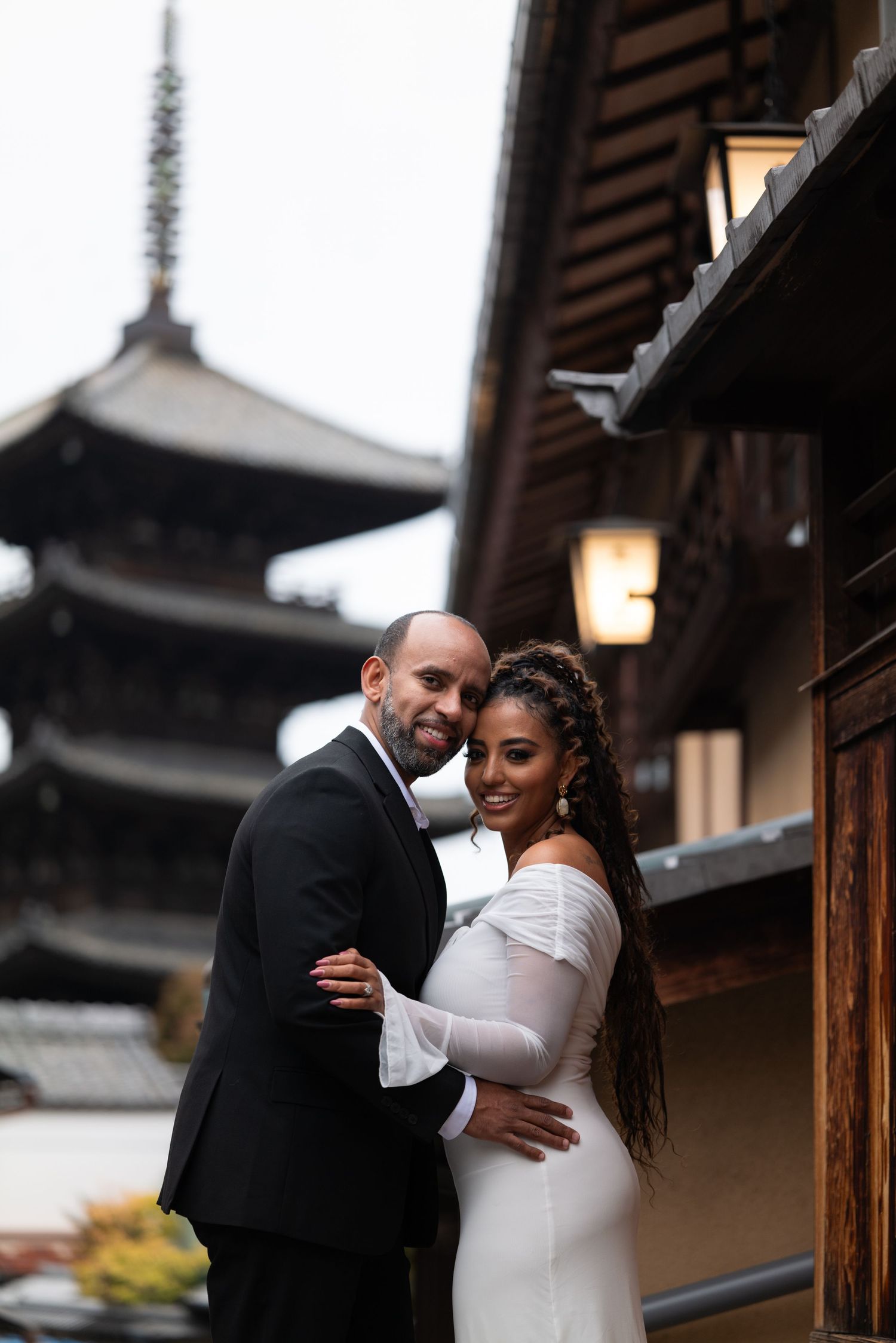 Couple embraces near Yasaka Pagoda in Kyoto, woman in white fitted gown, man in black suit.