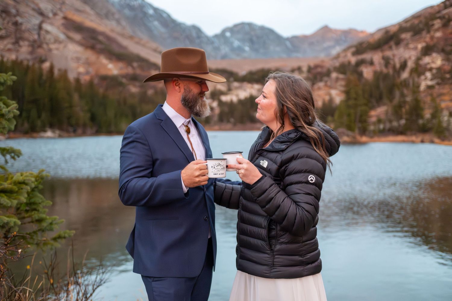 An eloping couple share coffee at sunrise by an alpine lake with dramatic mountain peaks behind them. She is in a black down coat.