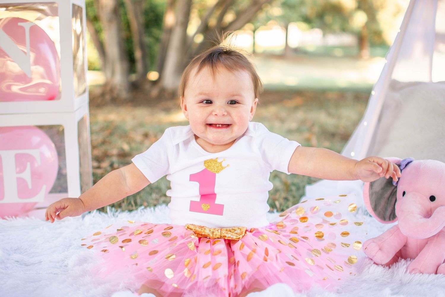 Baby wearing pink tulle skirt sits on white blanket with floral decorations during outdoor photo session.