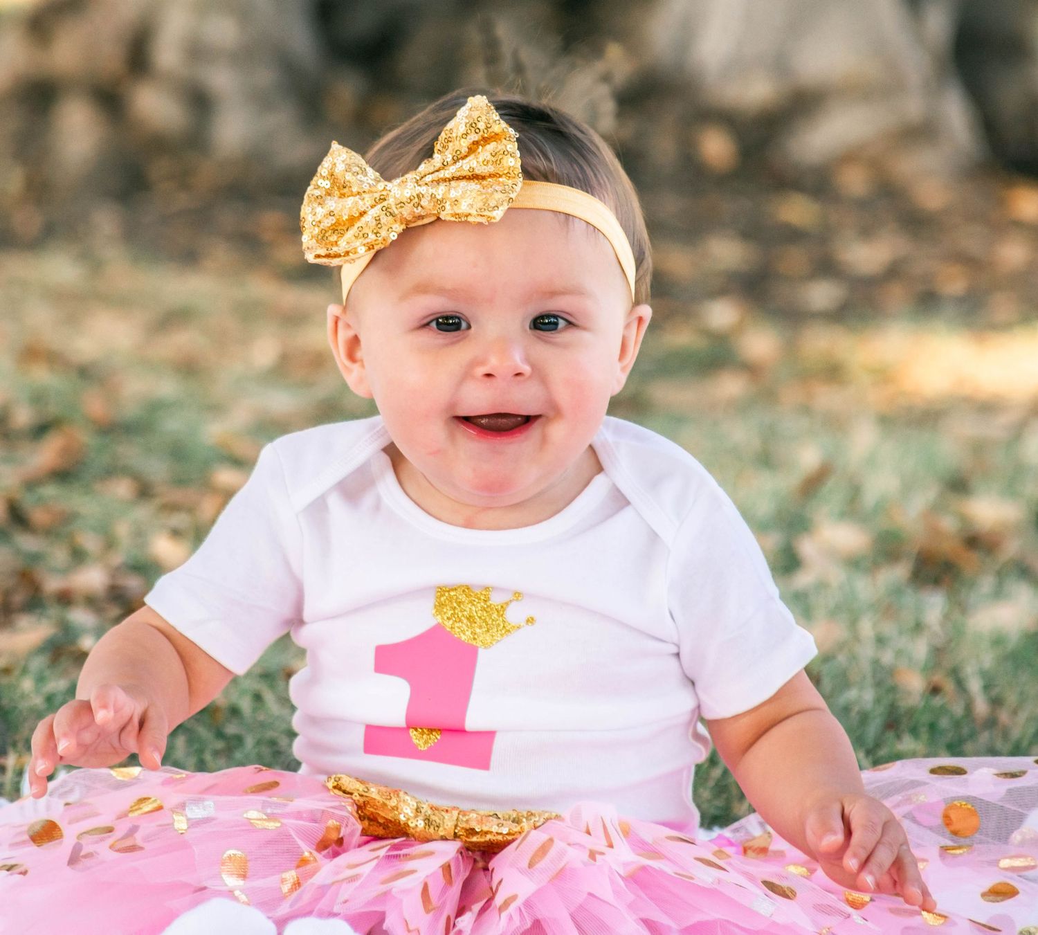Happy baby wearing a white shirt with number 1, gold bow headband and pink tutu sitting outdoors on autumn ground.