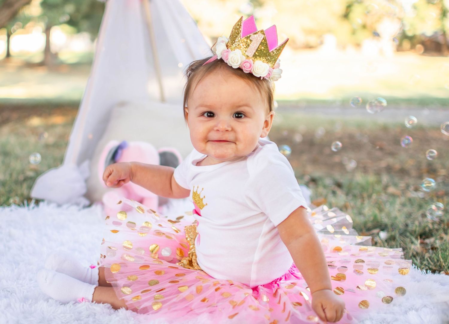 Baby in white outfit sits on pink tulle skirt surrounded by floral decorations in outdoor setting.