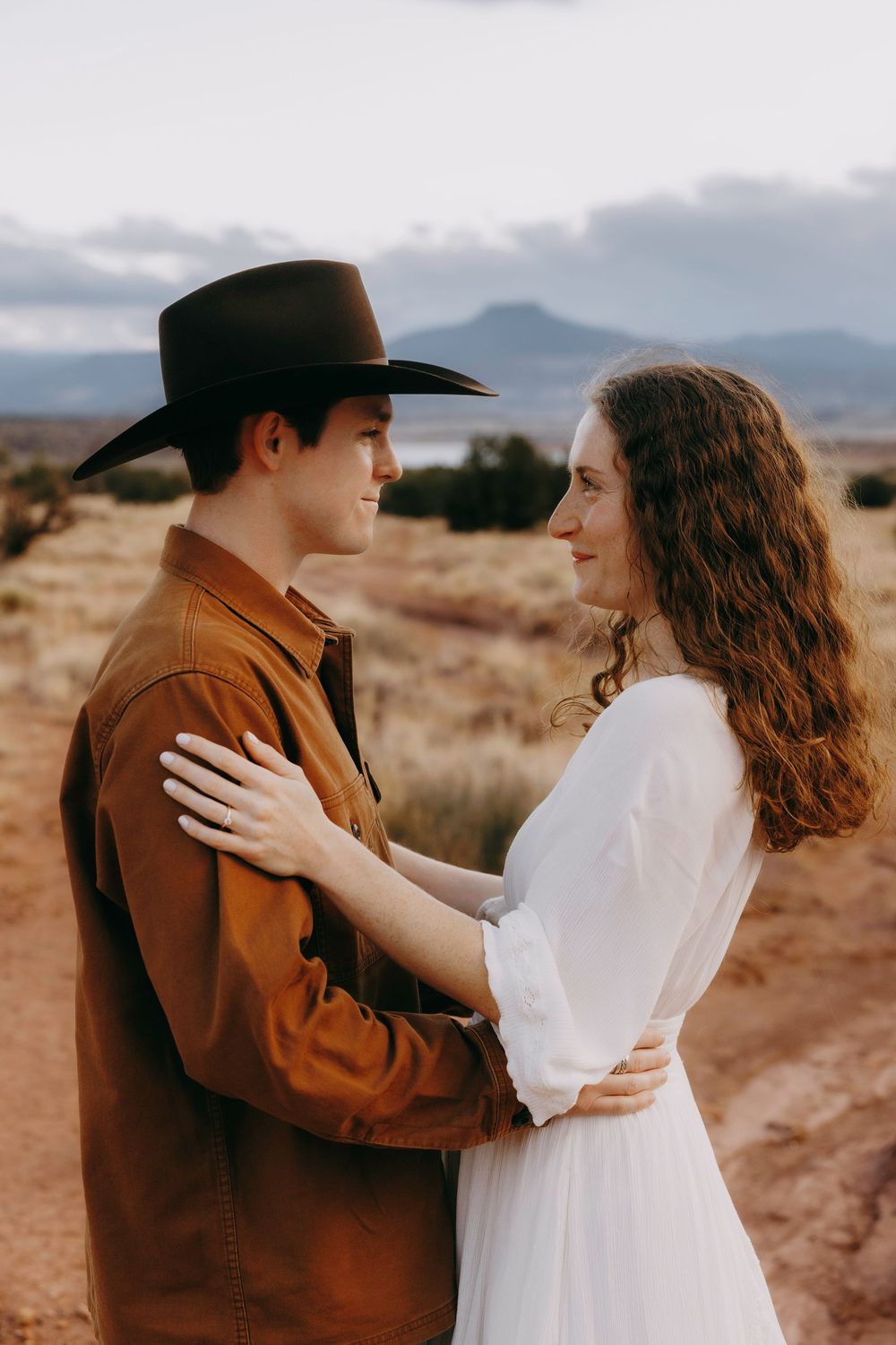Young man in cowboy hat and brown jacket embracing woman in white dress outdoors.