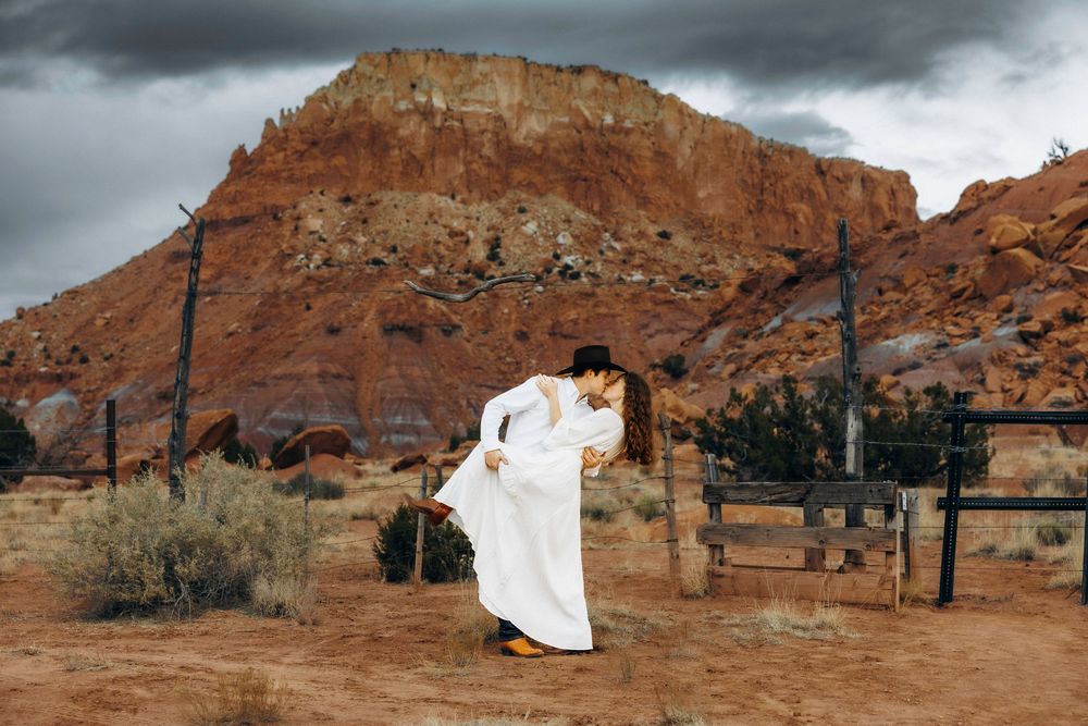Couple sharing romantic dip kiss in desert landscape with red rock formations and fence.