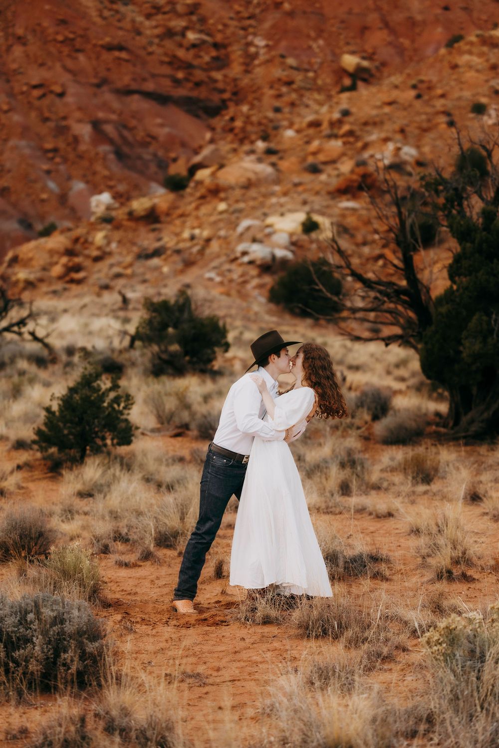 Couple kissing in desert landscape surrounded by red rock formations and desert shrubs.