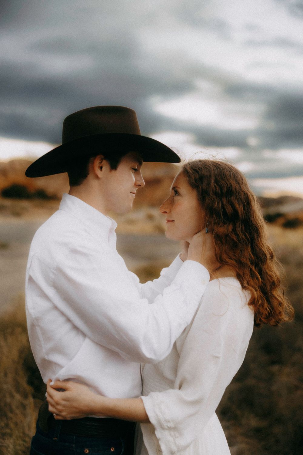 Man in black cowboy hat and white shirt embracing woman under dramatic stormy sky.