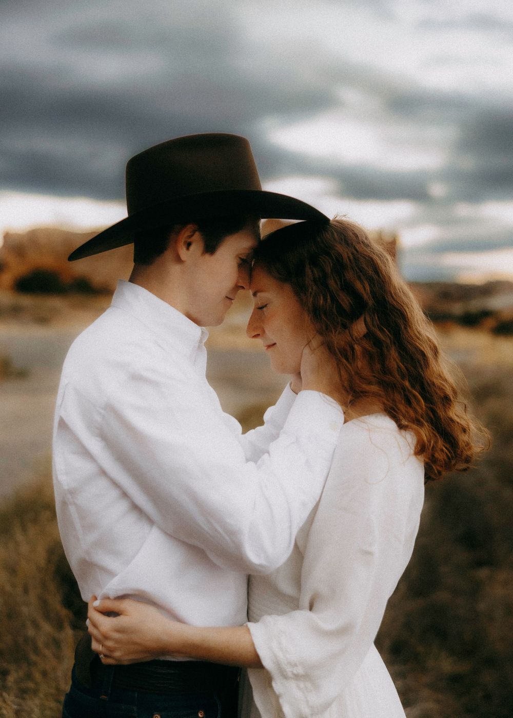 Couple in white shirts embrace forehead-to-forehead in a desert field under dramatic storm clouds, man wearing black cowboy hat.