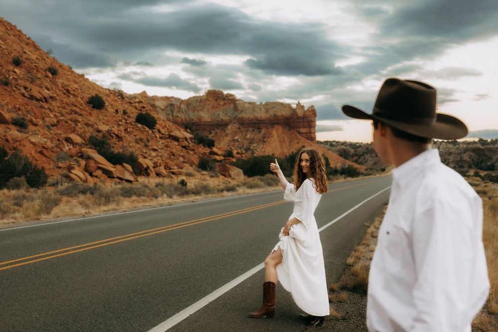 Woman in white dress and cowboy boots posing on desert highway with man in foreground.