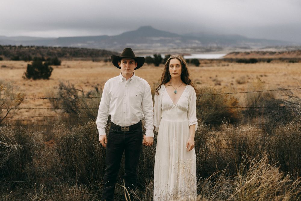 Santa Fe couple stands together in golden desert field, man in white shirt and cowboy hat, woman in white lace dress, mountains behind.
