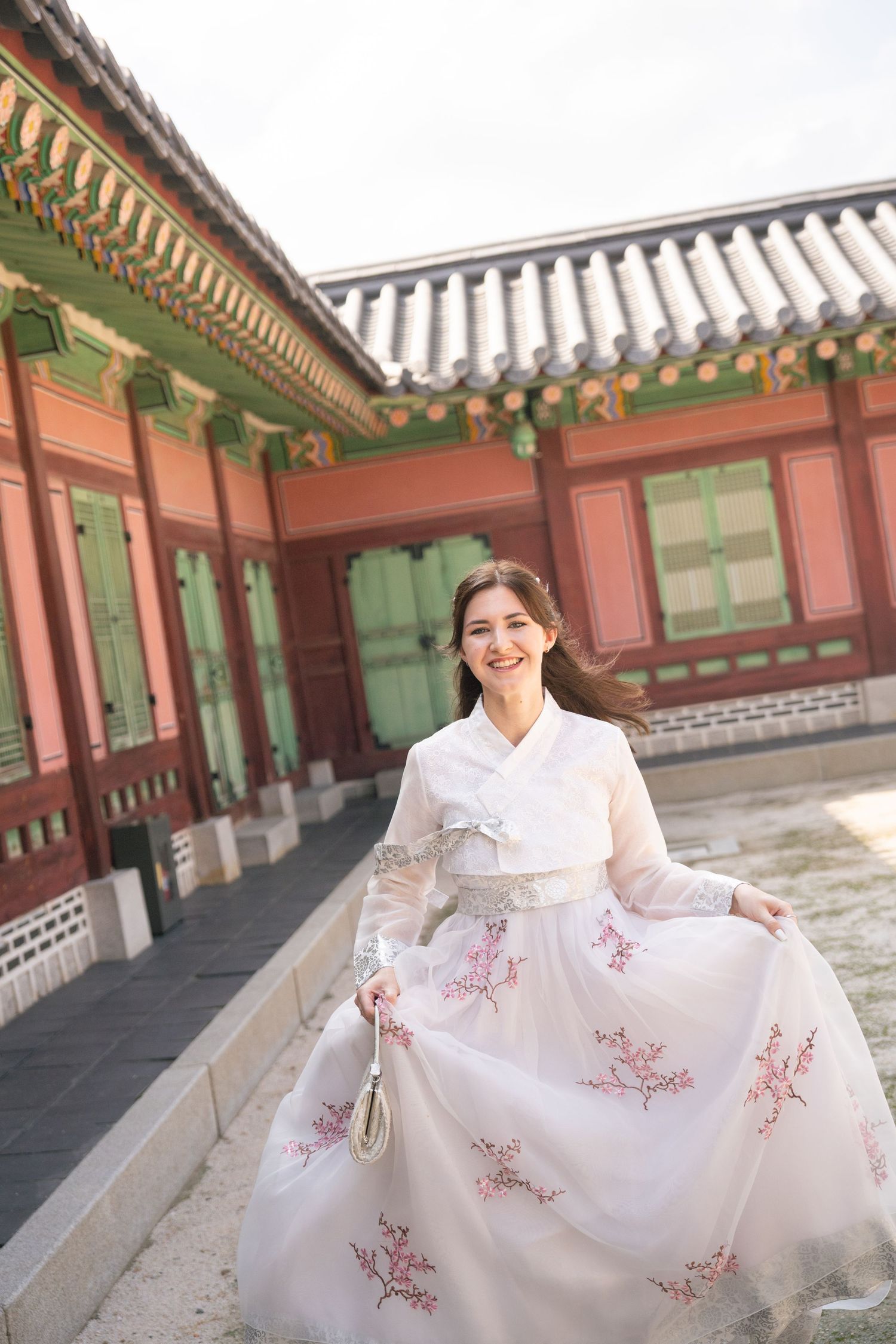 Woman wearing traditional Korean hanbok dress with floral embroidery stands in front of historic palace building.