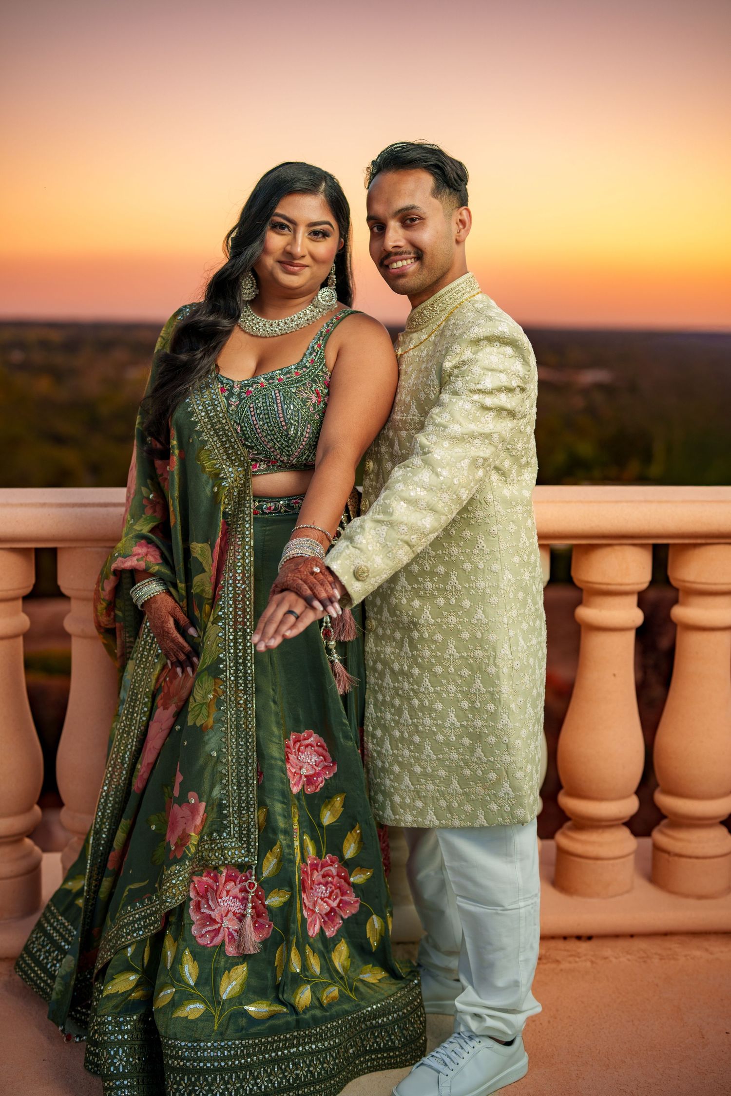 A couple in traditional Indian attire poses at sunset on a balcony with terracotta pillars, her emerald dress flowing elegantly.