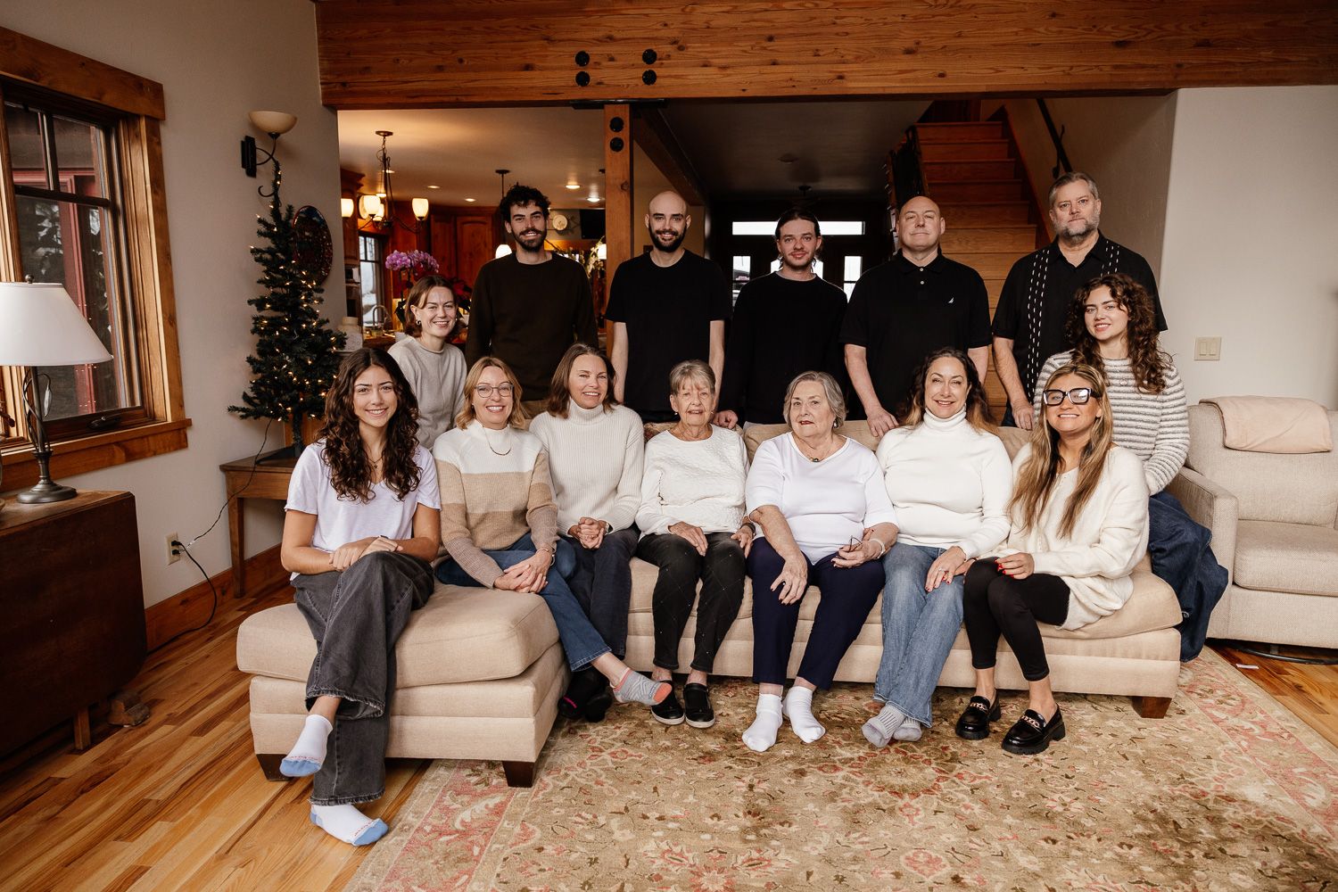 A large extended family group sits together on a beige couch in a cozy living room in their home with a Christmas tree in the background.