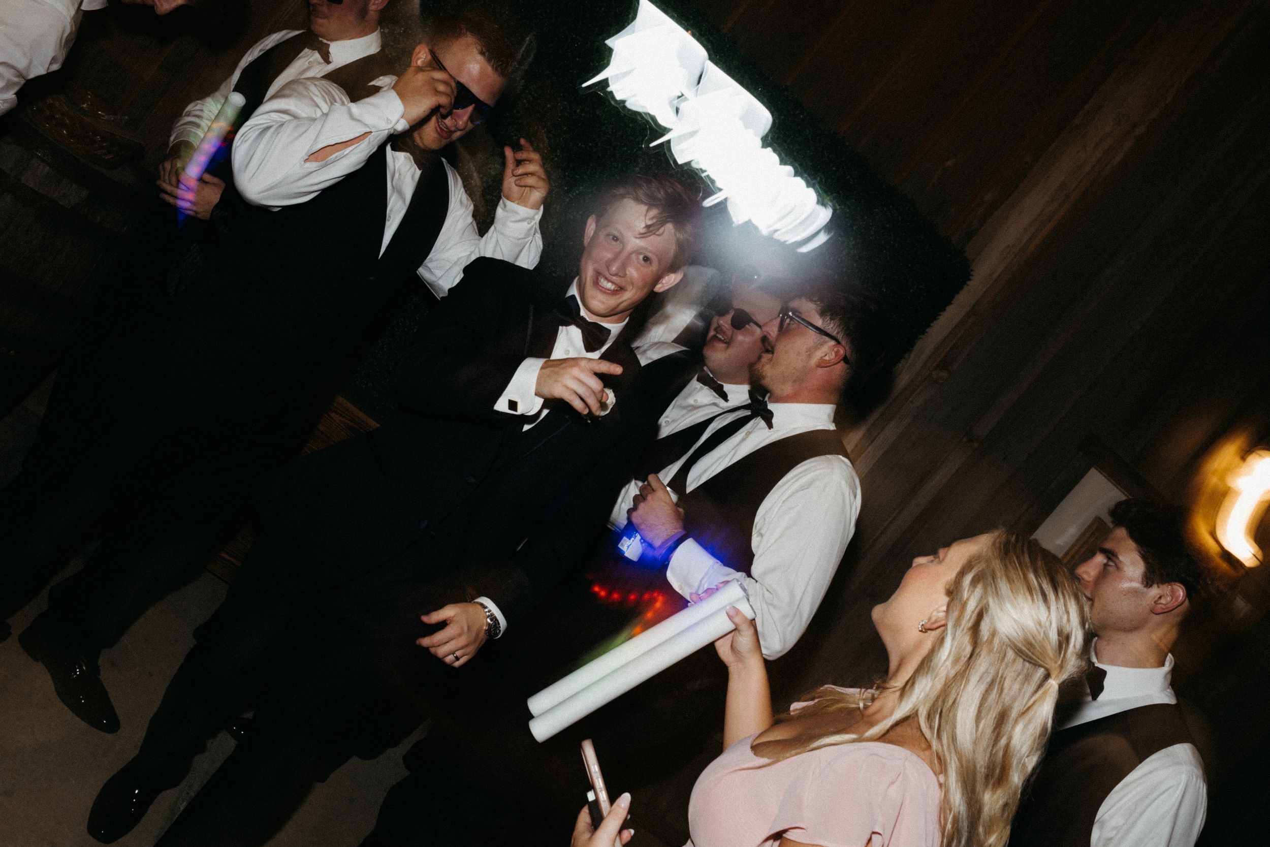 A group of people dancing and celebrating at a dimly lit party venue with colorful lights.