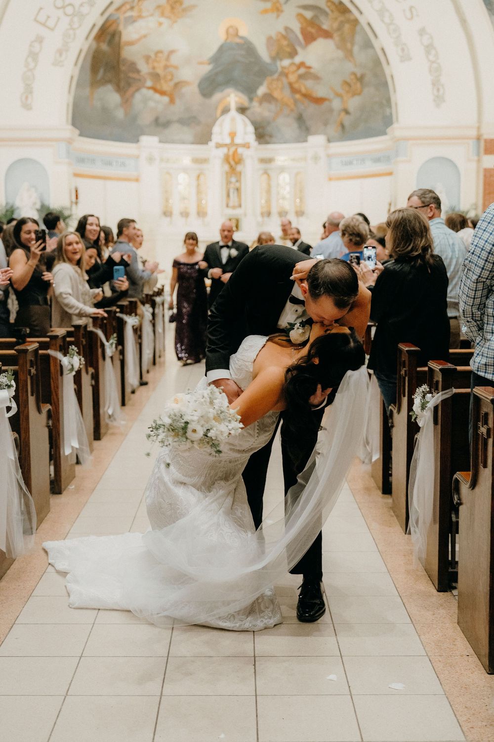 A couple shares a romantic dip kiss in a church aisle during their wedding ceremony as guests look on.