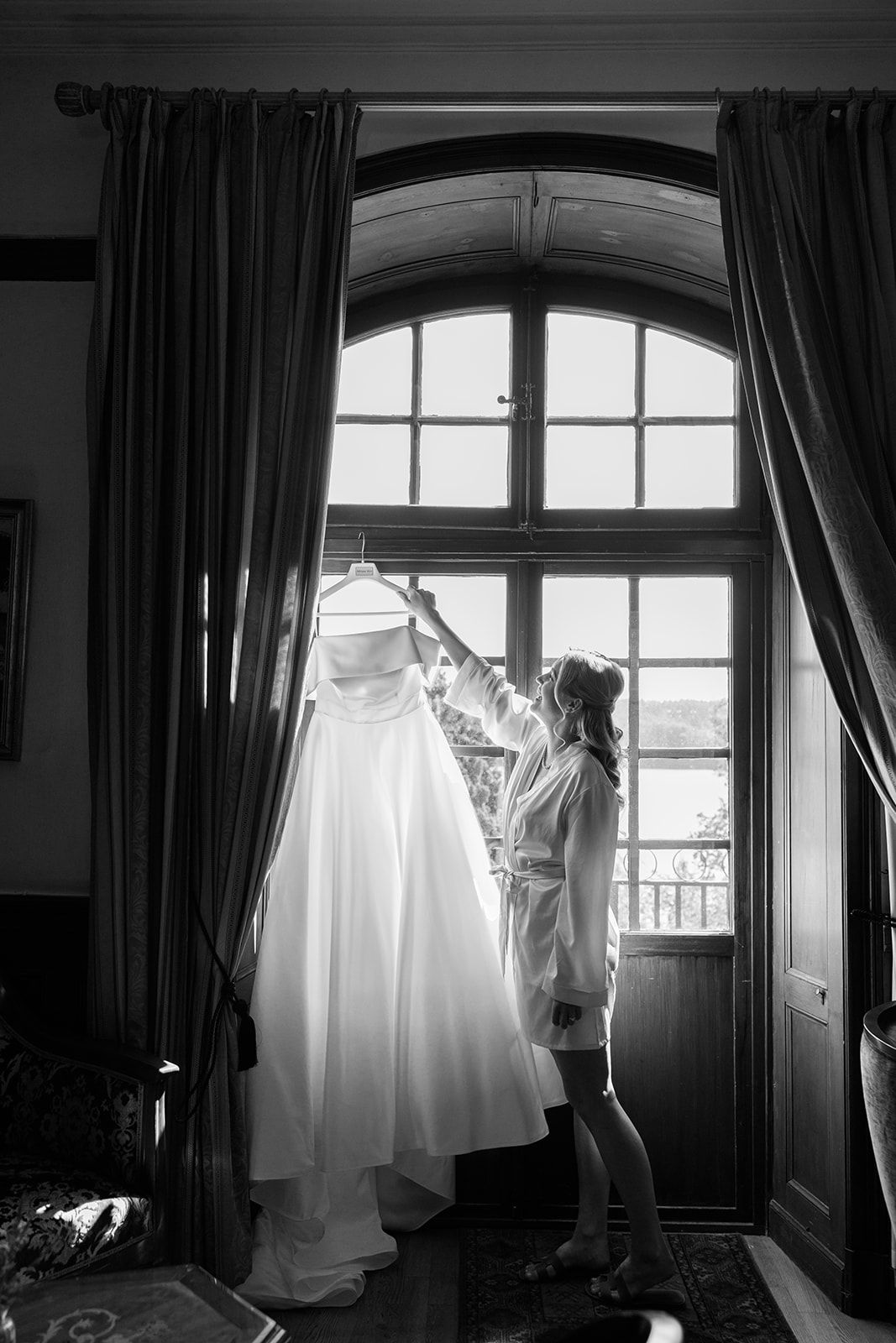 A wedding dress hangs in silhouette against a bright window with dark curtains in a dramatic black and white series.